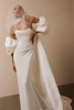 A model wearing a white satin bridal gown with dramatic puffed sleeves, a sheer veil, and a long train, posed against a plain beige wall.