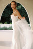 A woman modeling a sleeveless white wedding gown with thin straps and a flowing sheer overskirt, standing under a large arched doorway with greenery visible in the background.