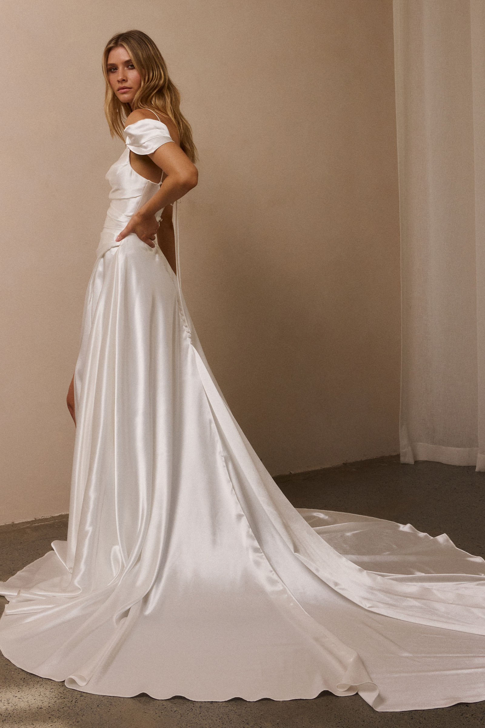 A woman modeling a white satin wedding gown with a short puff sleeve, a low back cutout, and a long train in a softly lit indoor setting_XS_