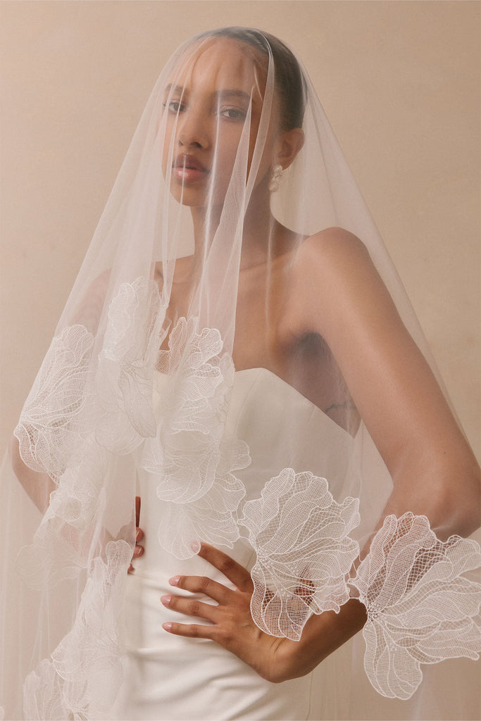 A woman wearing a long white bridal veil withembroidered lace and a strapless white dress, posed against a soft beige studio background.