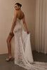 A woman modeling a strapless lace wedding gown from the back, showing a fitted lace bodice, high slit, sheer overlay, and long train in a softly lit indoor setting._XS_
