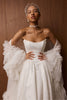 A woman modeling a strapless white wedding gown with dramatic ruffled sleeves, a sheer veil headpiece, and a jeweled choker against a warm wood-paneled background_XS