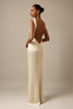A woman modeling a floor-length cream satin gown with a deep open back, shown from a rear three-quarter angle against a warm beige studio backdrop_XS_