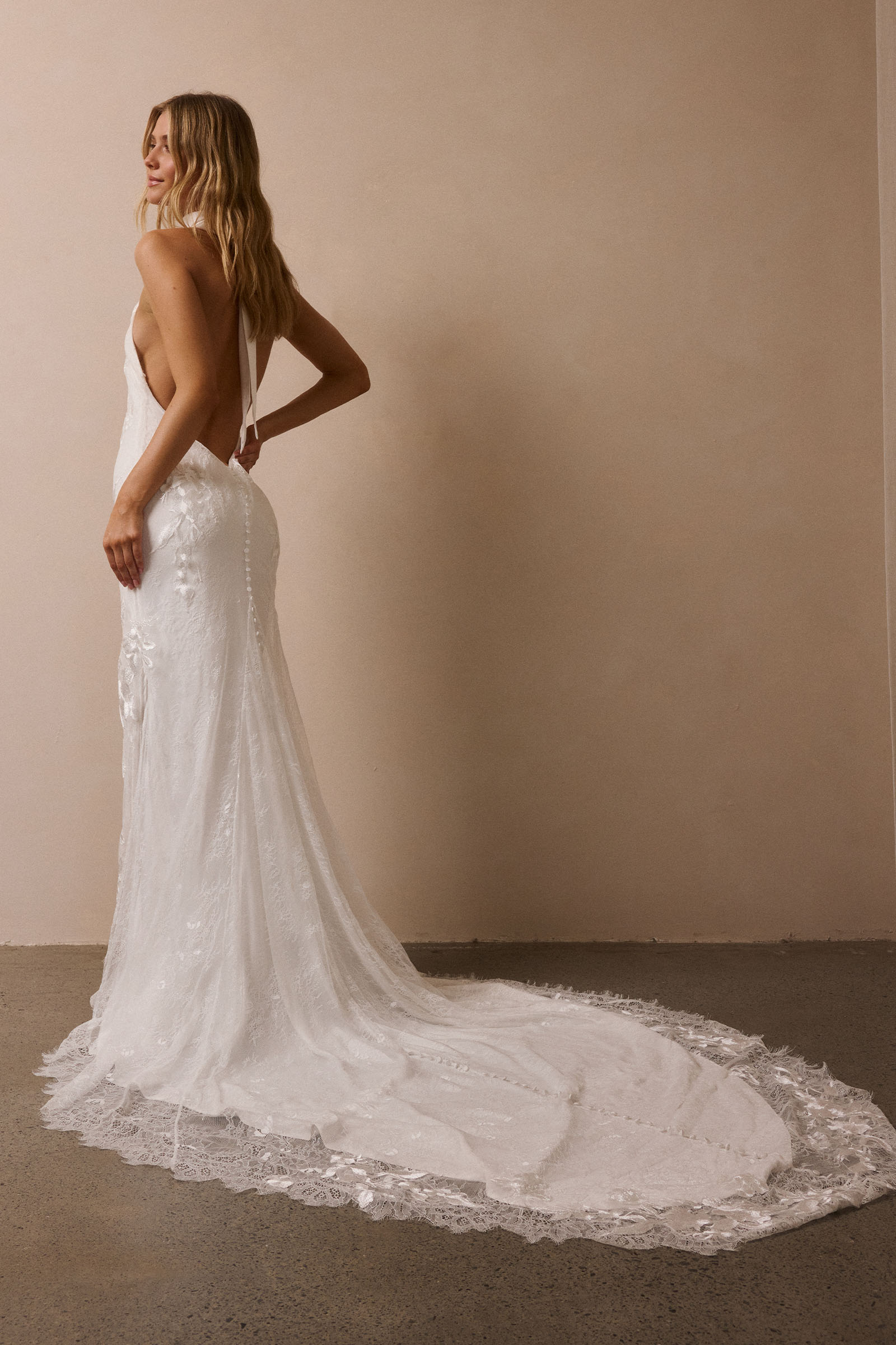 A woman modeling a white sleeveless wedding gown with a deep back and a long lace-trimmed train, standing against a plain beige wall_XS_