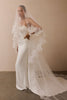 A woman modeling a strapless white wedding gown with a long sheer veil featuring floral lace applique, standing against a plain beige wall.