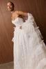 A woman modeling a strapless white wedding gown with a fitted lace bodice, layered tulle overskirt, and sheer draped sleeves, posed against a warm wood-paneled wall.