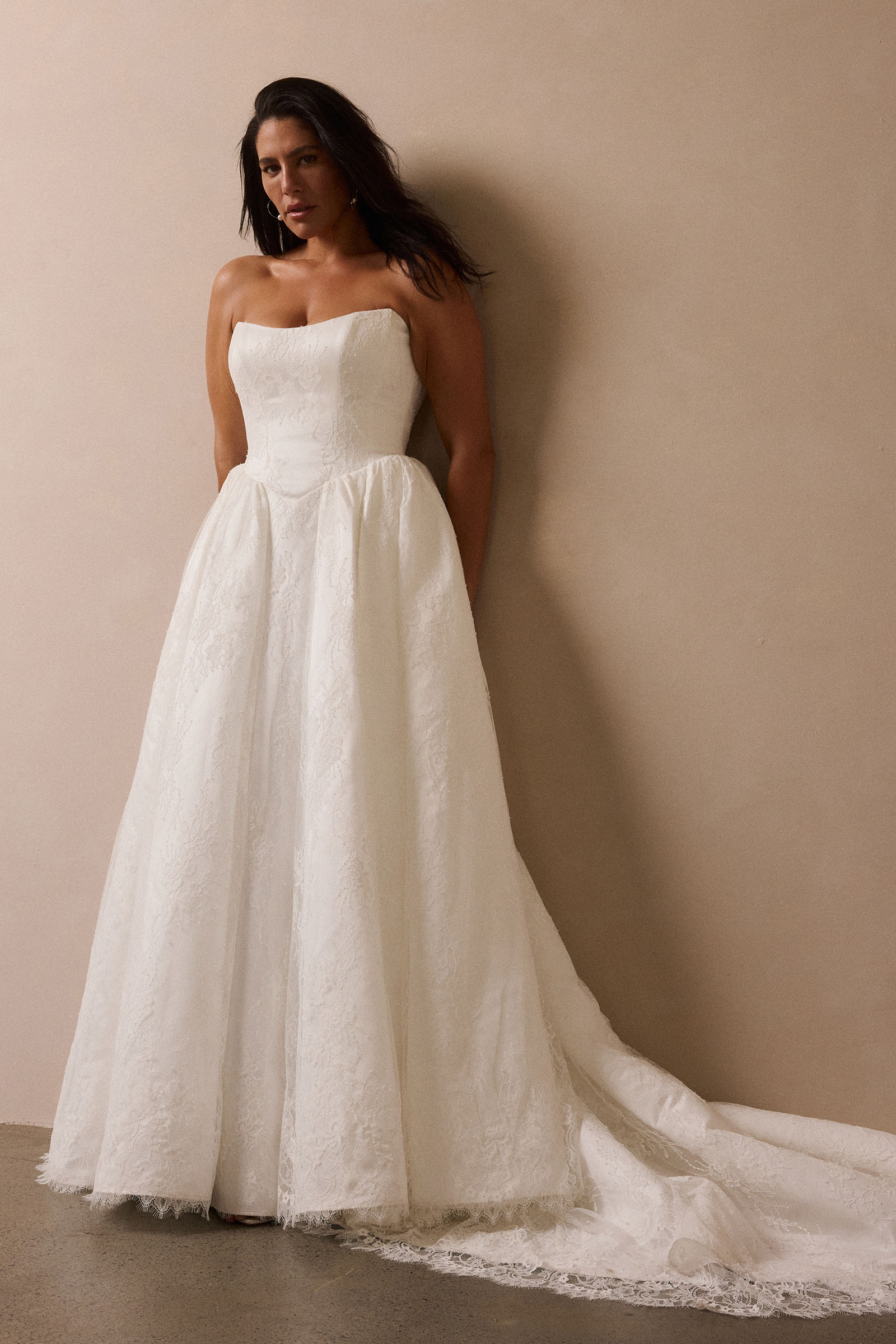 A woman wearing a strapless white wedding gown with a fitted bodice and full lace-trimmed skirt, standing against a plain beige wall_L_
