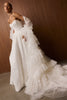 A woman modeling a dramatic white wedding gown with a strapless bodice, sheer long sleeves, and a layered ruffled overskirt and train, posed against a dark wood-paneled wall_XS