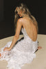 A woman seated on a circular platform wearing a white lace backless dress with thin straps and delicate chain details across the open back, photographed against a dark studio background.