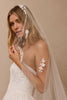A woman wearing a white bridal dress and a sheer white veil decorated with floral lace appliques, posed against a warm beige studio background.