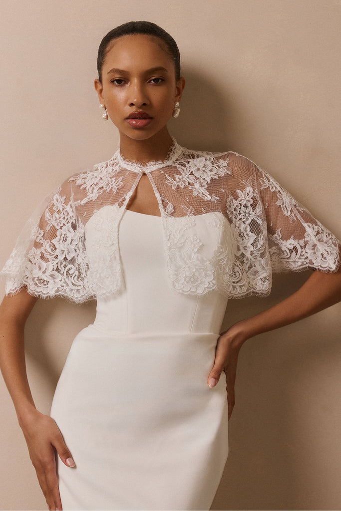 A woman modeling a white sleeveless dress with a sheer white lace bolero-style shrug over the shoulders, posed against a plain beige studio wall.