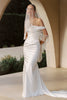 A woman wearing a white off-the-shoulder mermaid-style gown with long fringe draped over the arms and a matching veil, standing in a sunlit architectural setting with arches and greenery in the background.