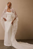A woman modeling a white strapless wedding gown with a long train and a sheer lace veil draped over her shoulders, posed against a plain beige studio wall.