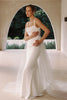 A woman modeling a fitted white wedding dress with thin straps, a draped neckline, and a long sheer tulle overskirt, posed in front of an arched architectural opening.