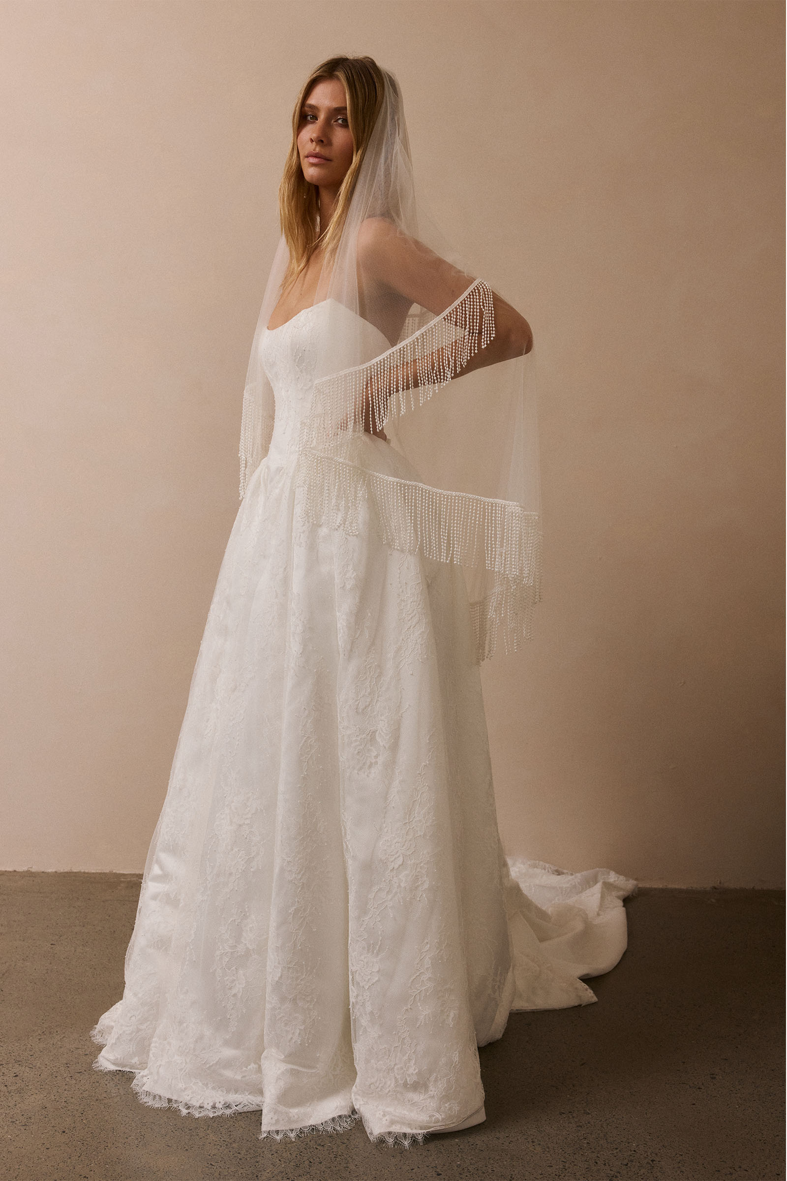 A woman wearing a white lace bridal gown with a long sheer veil and fringe detail, standing in a studio with a warm beige wall and dark floor_XS_