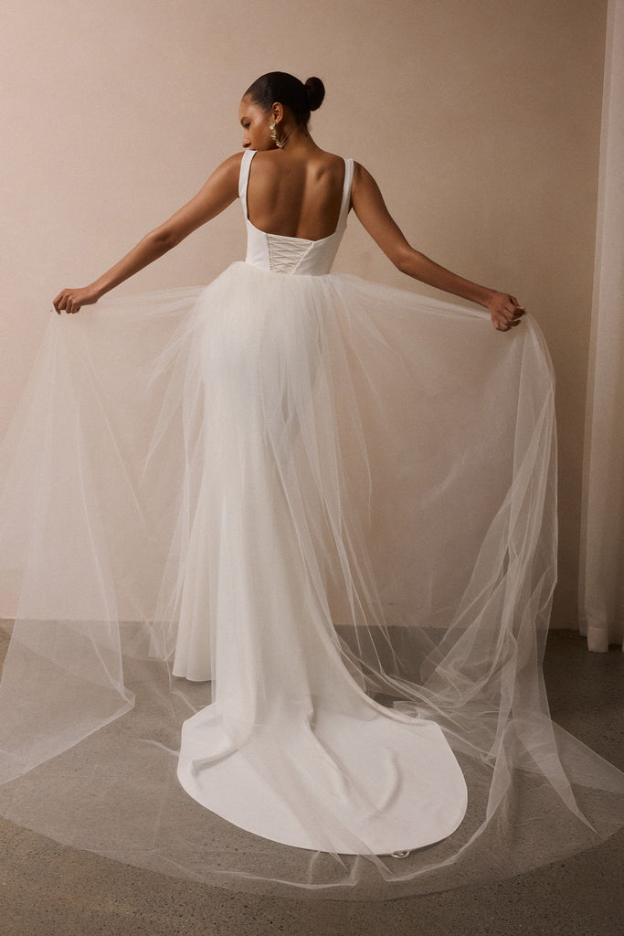 A woman stands with her back to the camera wearing a white wedding dress with a structured backless bodice and a very full tulle overskirt and train, holding the fabric out with both hands in a studio with a beige wall and gray floor.