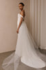 A woman modeling a sleeveless white wedding gown with a fitted bodice, full skirt, and long sheer tulle train against  a plain beige studio wall_XS_