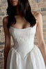 A close-up of a woman wearing a strapless white lace corset-style dress with a full skirt, standing against a rustic brick wall.