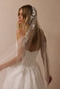 A woman wearing a white wedding dress and a sheer white bridal veil with floral lace applique, photographed against a soft beige studio background.