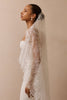 A woman in profile wearing a white wedding dress and a sheer lace bridal veil against a plain beige studio background.