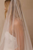 A woman in profile wearing a long, sheer white bridal veil over her hair and shoulders, with part of a white lace wedding dress visible against a soft beige background.