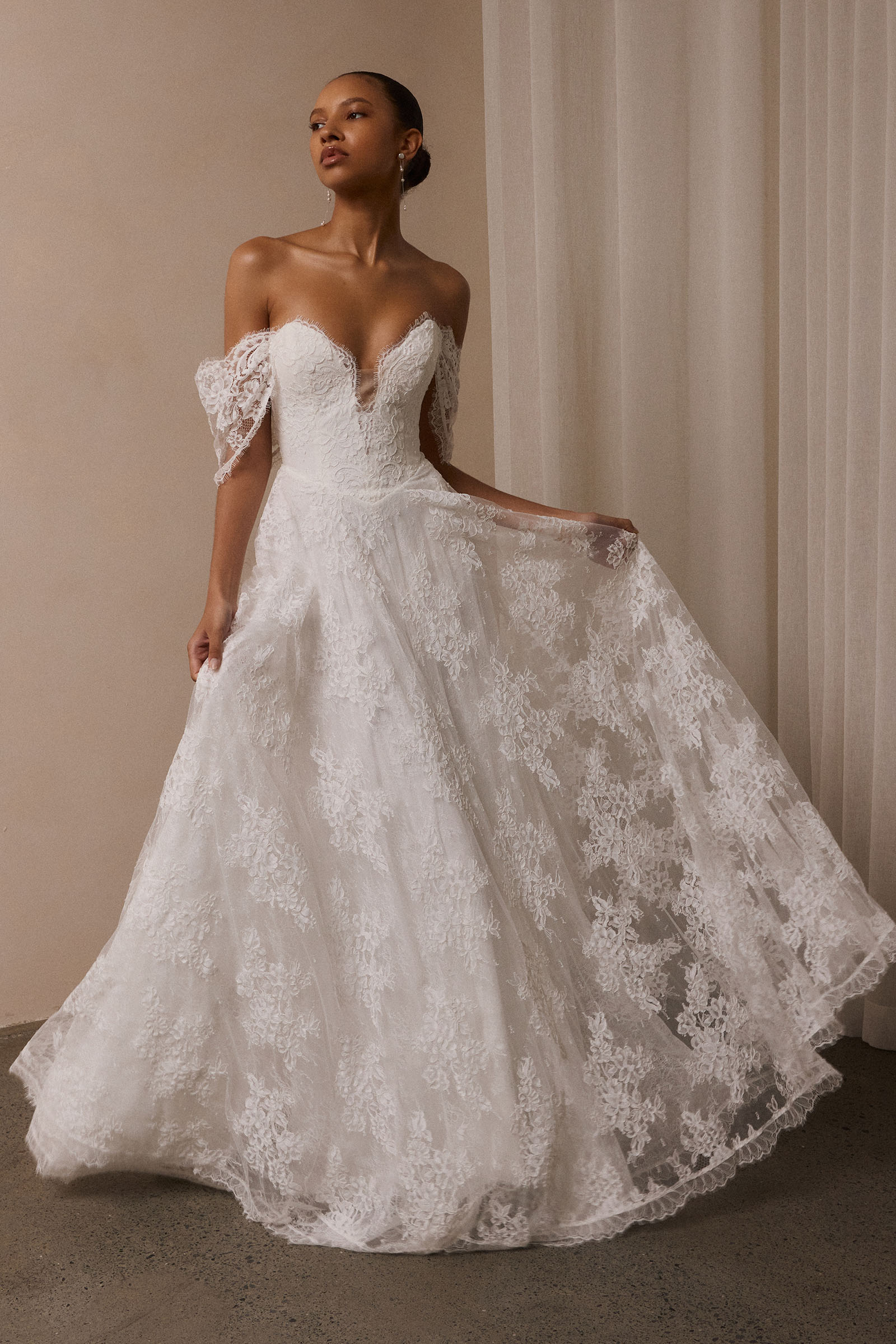 A woman wearing a white off-the-shoulder lace wedding gown with a deep V neckline and a full layered skirt_XS_
