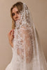 A woman wearing a white lace bridal veil over her face and shoulders, with a white wedding dress visible at the bottom, posed against a plain beige studio background.