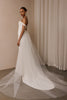 A woman modeling a white off-the-shoulder wedding gown with a long sheer tulle train in a softly lit indoor setting with beige walls and draped fabric.