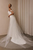 An off-the-shoulder white wedding gown with a long sheer tulle train, modeled in a softly lit indoor setting with beige walls and draped curtains.