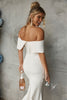 Woman in off-shoulder wedding dress holding a bottle and glass against a gray wall.