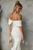 Woman in off-shoulder wedding dress holding a bottle and glass against a gray wall.